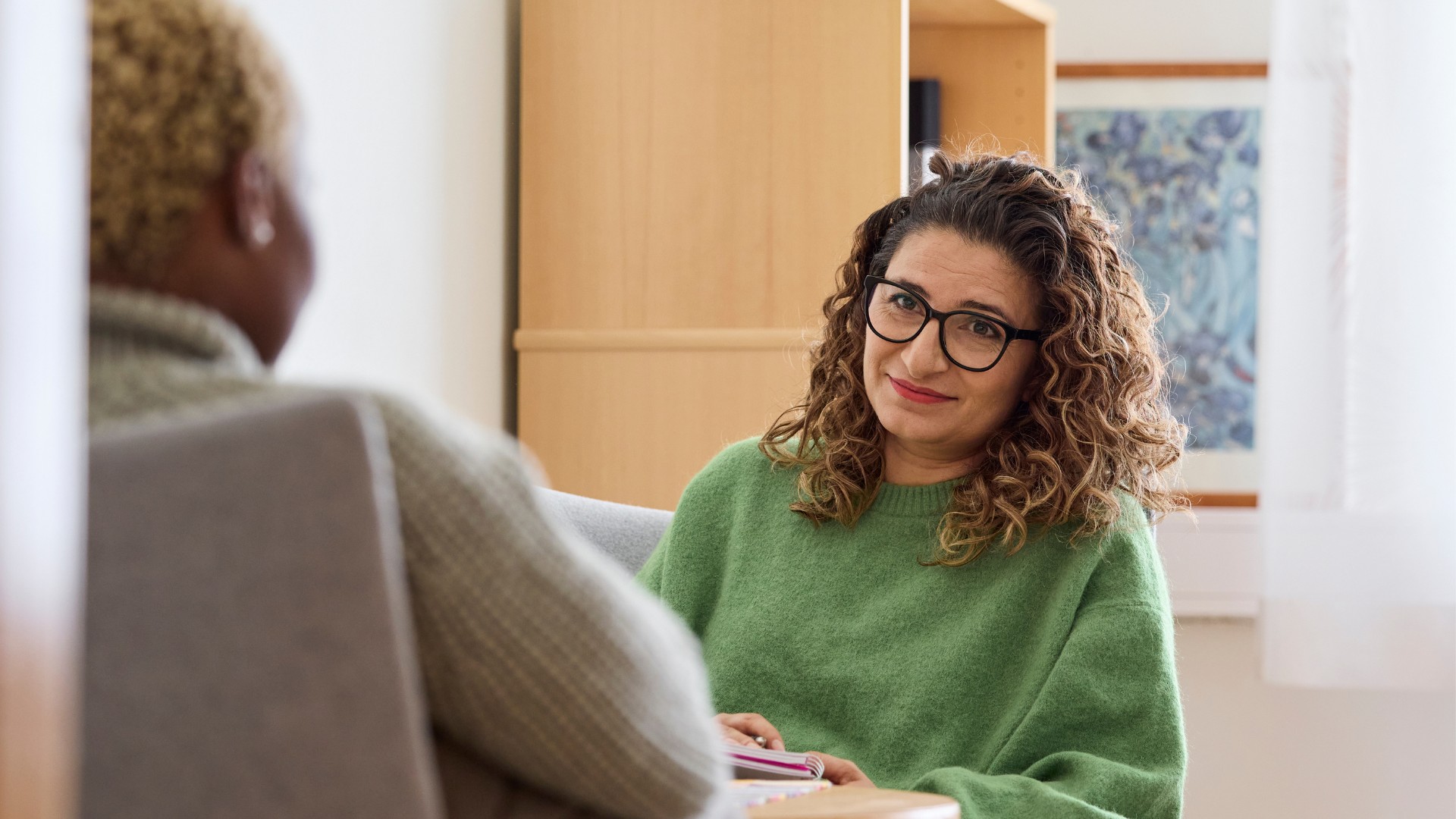 Två personer sitter mittemot varandra i ett samtal. Den ena personen, som bär grön tröja och glasögon, tittar vänligt mot kameran. Den andra personen syns bakifrån. Miljön är varm och lugn, som vid ett stöd- eller terapitillfälle.
