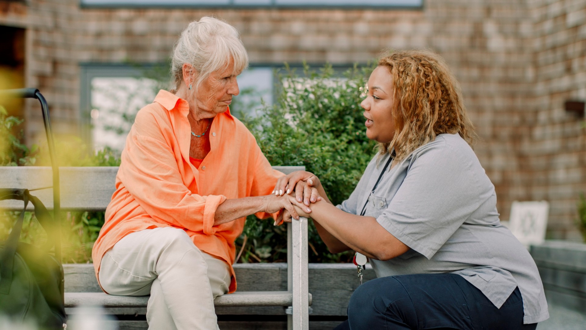En äldre kvinna sitter på en bänk utomhus och håller händerna på en yngre kvinnas, som sitter framför henne i samtal. De ser varandra i ögonen och delar ett stöttande ögonblick. Miljön är grön och rofylld.