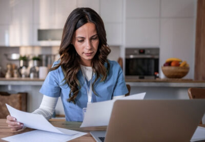 Person inom vård och omsorg som läser dokument och studerar vid laptop i hemmiljö.