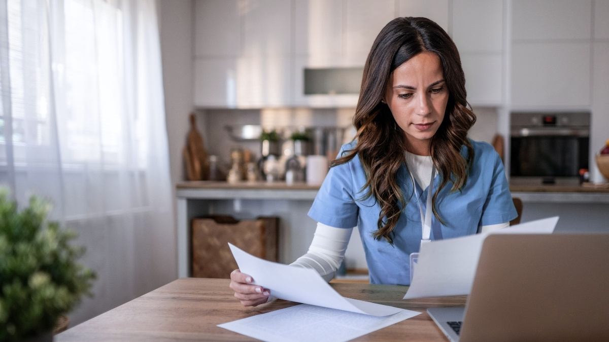 Person i vårdkläder som studerar vid laptop hemma och går igenom dokument – distansstudier inom vård och omsorg.