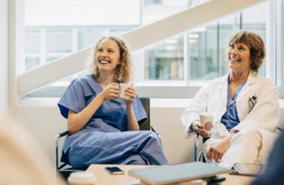 En ung kvinna med lockigt blont hår och en äldre kvinna med kort brunt hår sitter tillsammans och ler glatt, båda i arbetskläder. Den unga kvinnan håller en kopp kaffe i händerna och ser ut att skratta, medan den äldre kvinnan håller en pappersmugg. De sitter i en ljus och modern miljö med stora fönster som släpper in mycket naturligt ljus. Omsorg och återhämtning i vården. Vårdmedarbetare tar en paus i lugn miljö.