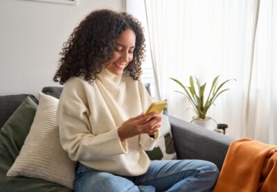 Leende person sitter bekvämt i en soffa hemma, tittar ner på sin mobiltelefon. Ljust rum med kuddar, filt och en grön växt i bakgrunden.