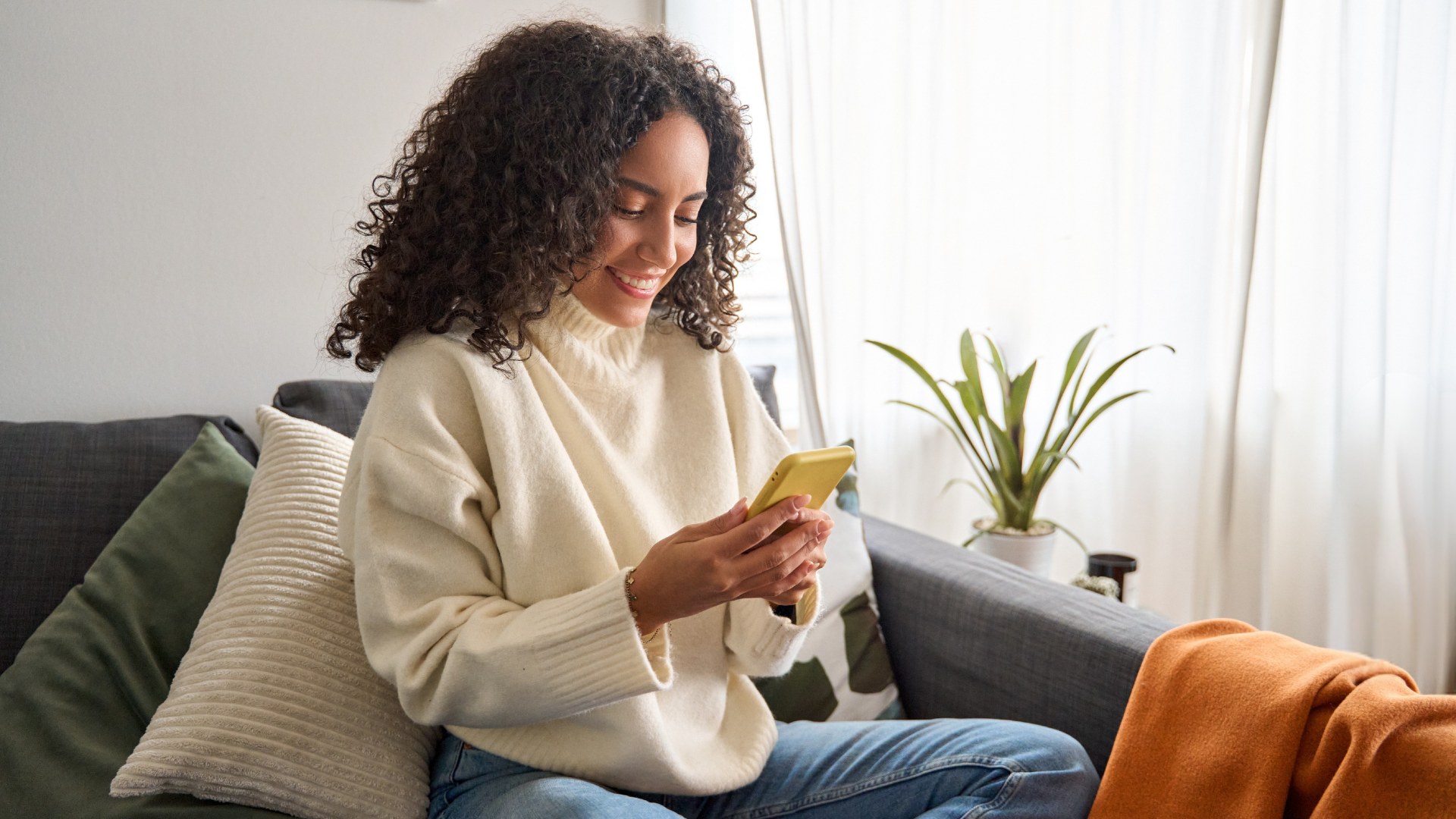Leende person sitter bekvämt i en soffa hemma, tittar ner på sin mobiltelefon. Ljust rum med kuddar, filt och en grön växt i bakgrunden.