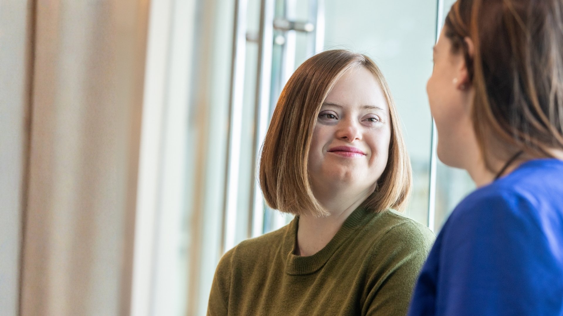 Person med funktionsnedsättning sitter i samtal med vårdpersonal. De har ögonkontakt och möts i en lugn och respektfull dialog. Bilden illustrerar delaktighet, självbestämmande och ett inkluderande bemötande.