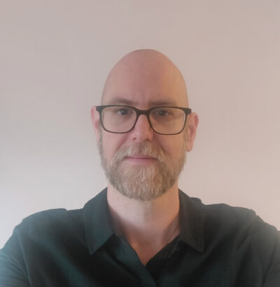 Porträttfoto av en man (Alois) med glasögon, skägg och rakad hjässa. Han bär en mörkgrön skjorta och tittar rakt in i kameran med ett neutralt uttryck. Bakgrunden är ljus och enfärgad.