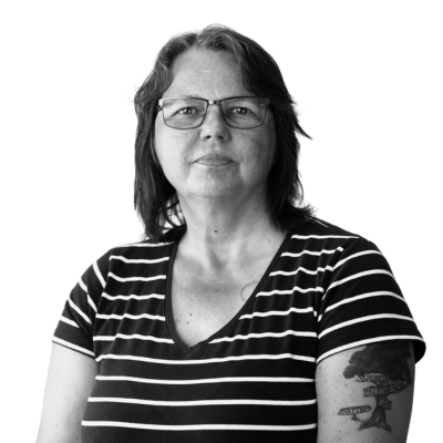 Porträttfoto i svartvitt av Tina Silkhage, utbildningsledare. En kvinna med glasögon, mörkt axellångt hår och randig tröja, som ser rakt in i kameran.