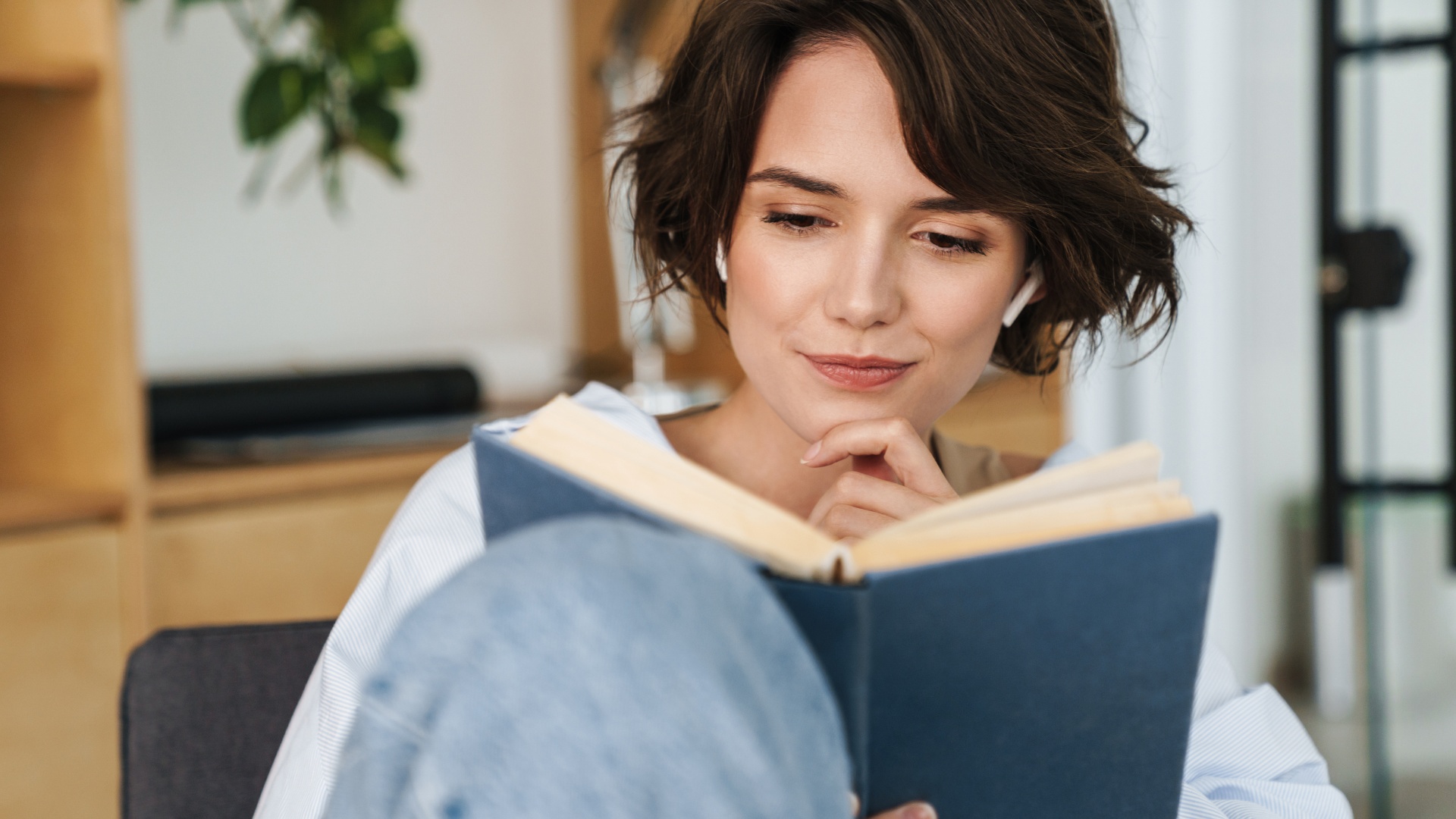 Ung kvinna med kort mörkt hår och trådlösa hörlurar som sitter avslappnat hemma och läser en bok, med fokus och en koncentrerad blick.
