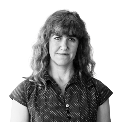 Porträttfoto i svartvitt av Josefin Kärling Elofsson, utbildningsledare. En kvinna med lockigt hår och lugg, klädd i en mörkprickig blus, som ser rakt in i kameran.