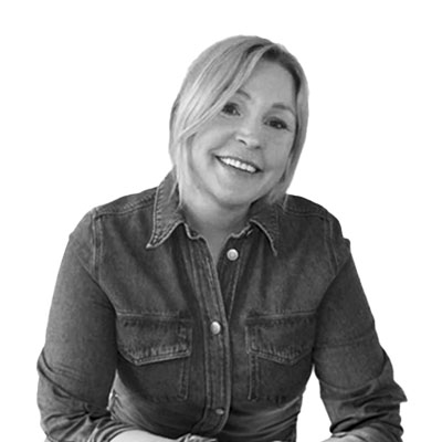 En porträttbild på Marie Schulz, utbildningsledare på Vårdyrkeshögskolan. En svartvit bild där hon sitter och ler. Hon har på sig en jeansskjorta med knappar och har en avslappnad hållning mot en vit bakgrund.