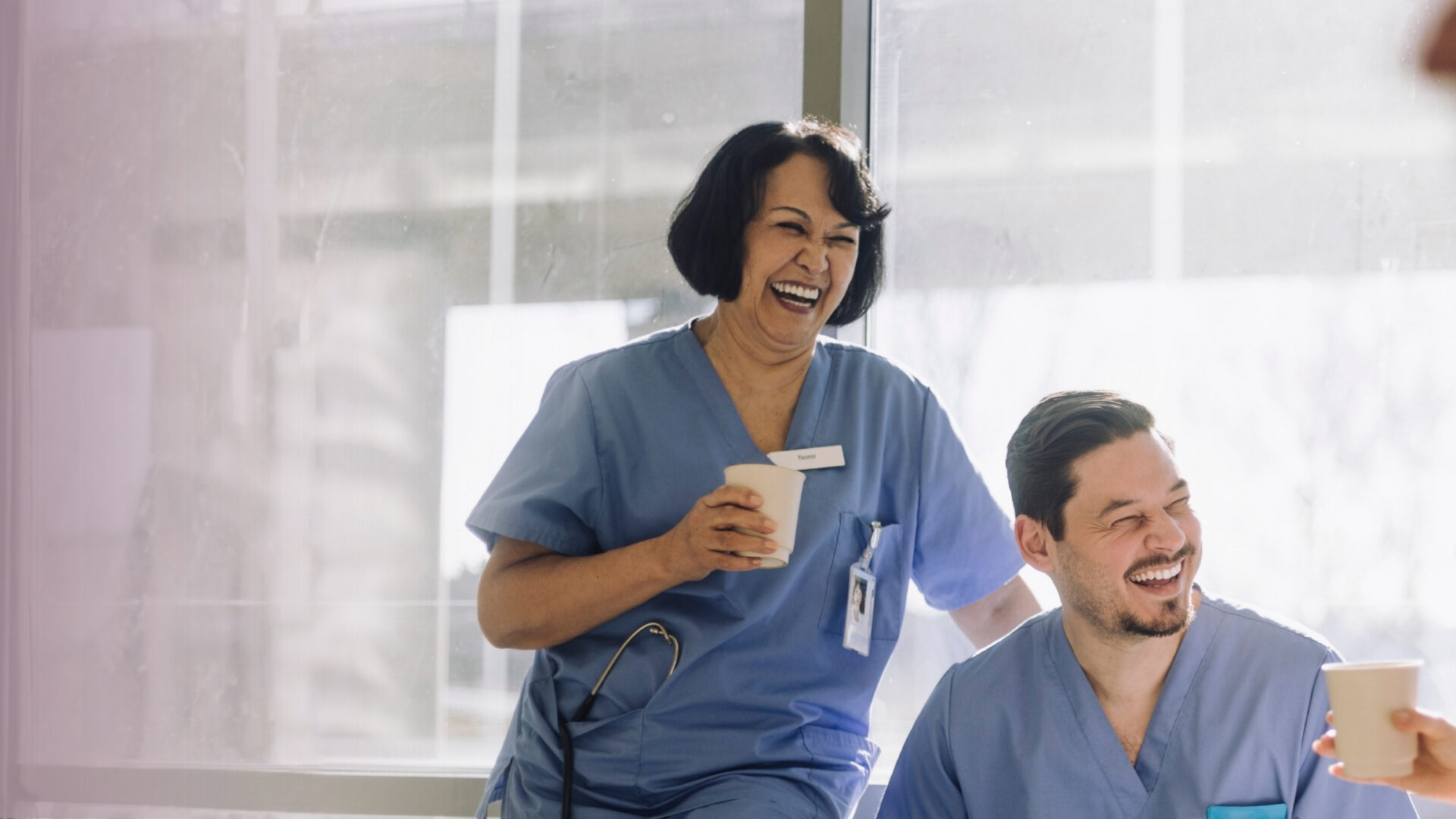 Tre personer i vårdkläder sitter tillsammans och skrattar under en paus. Bilden visar gemenskap och arbetsvardag inom vård och omsorg.